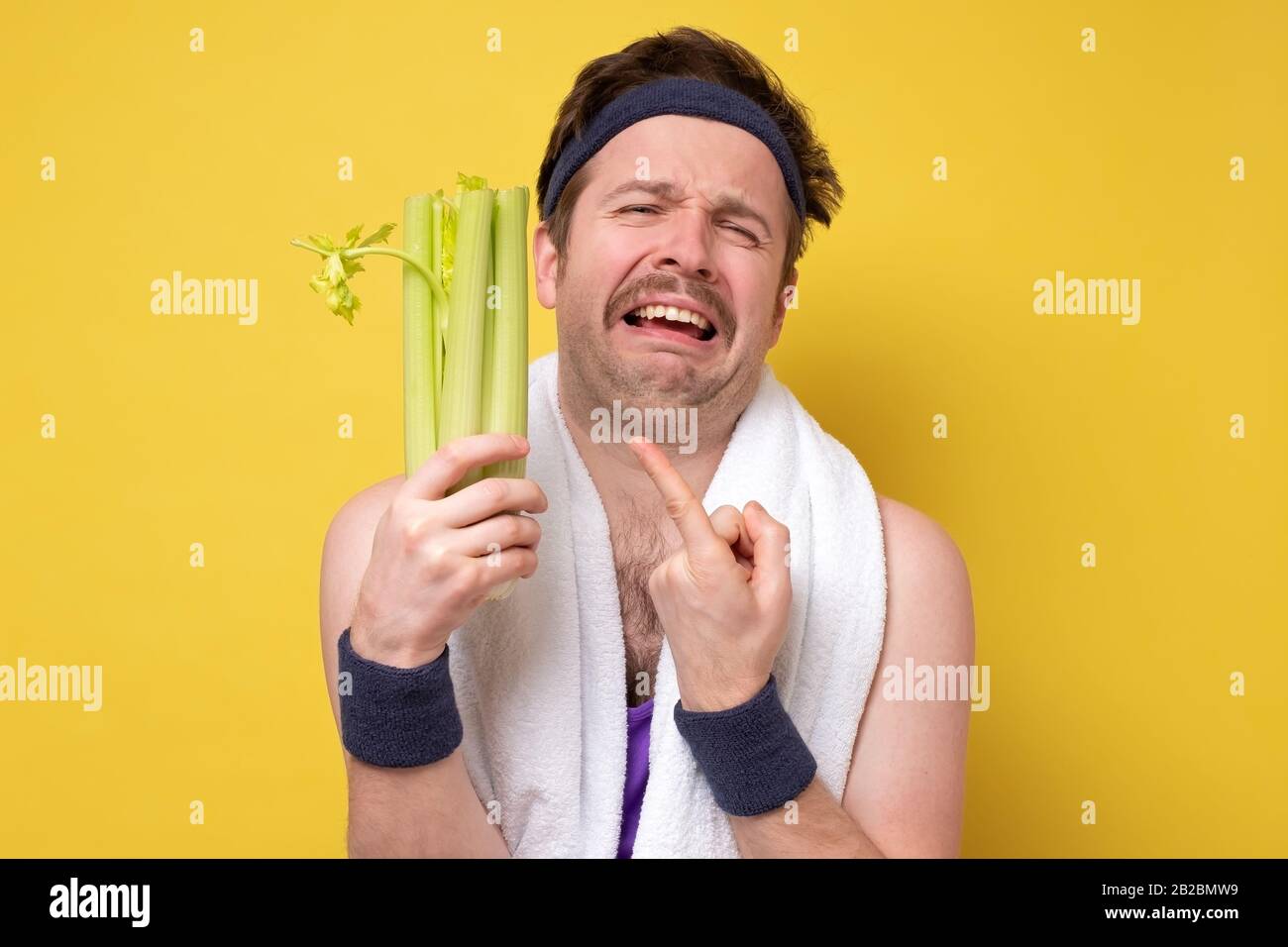 Lustiger sportlicher Mann mit Schnurrbart, der frischen Sellerie weinend hält. Ich hasse Gemüse, sollte aber für eine gesunde Ernährung essen. Studio an gelber Wand gedreht Stockfoto