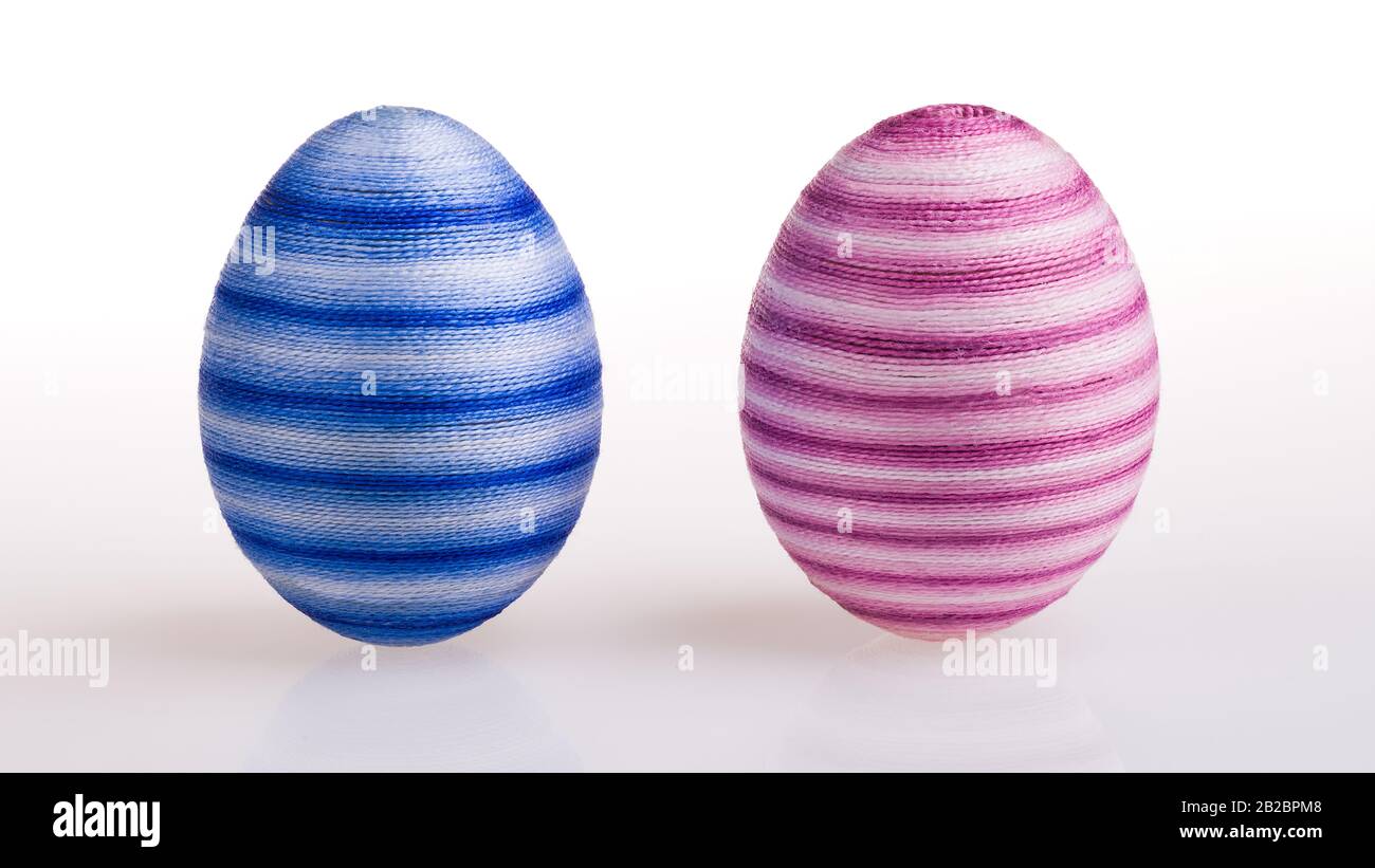 Zwei stehende Ostereier gestreift in männlicher blauer und weiblicher rosafarbener Farbe auf weißem Grund. Farbige, gestreute Eierschalen, die von geklebten dünnen Baumwollgarnen umhüllt werden. Stockfoto