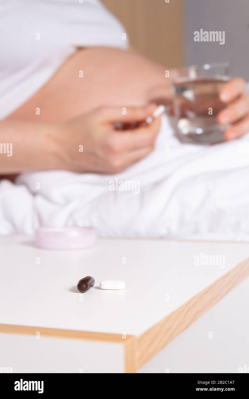 Unerkennbare Schwangere Frau im Bett, die Vitamine, Pillen in der Hand vor der Aufnahme hält. Glas Wasser in den Händen. Schwangerschaft gesunder Lebensstil. Schneeweiß B. Stockfoto