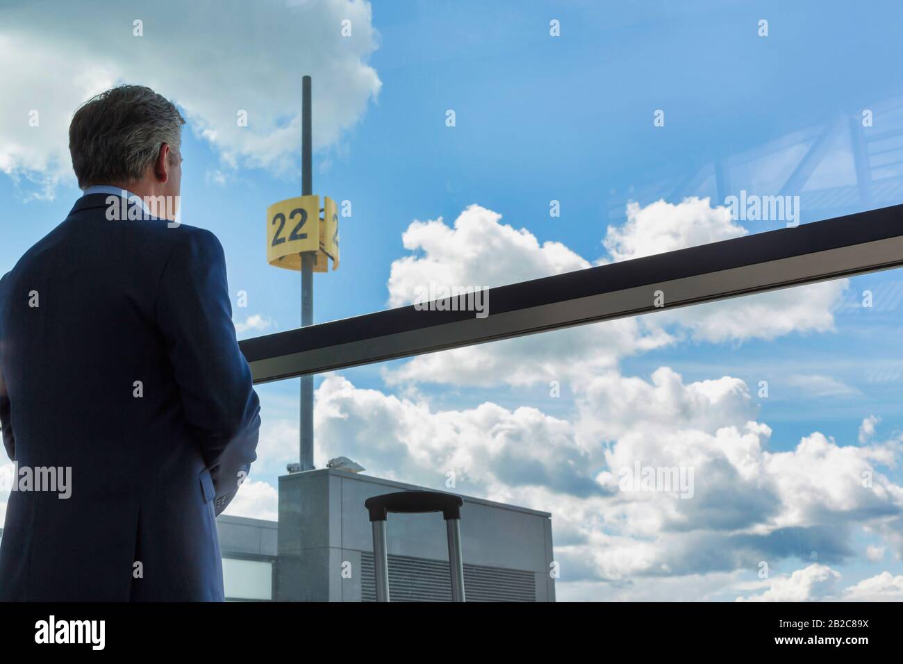 Porträt eines reifen Geschäftsmannes, der mit seinem Koffer steht, während er durch das Fenster des Flughafens blickt Stockfoto