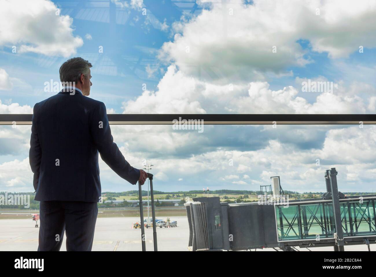 Porträt eines reifen Geschäftsmannes, der mit seinem Koffer steht, während er durch das Fenster des Flughafens blickt Stockfoto
