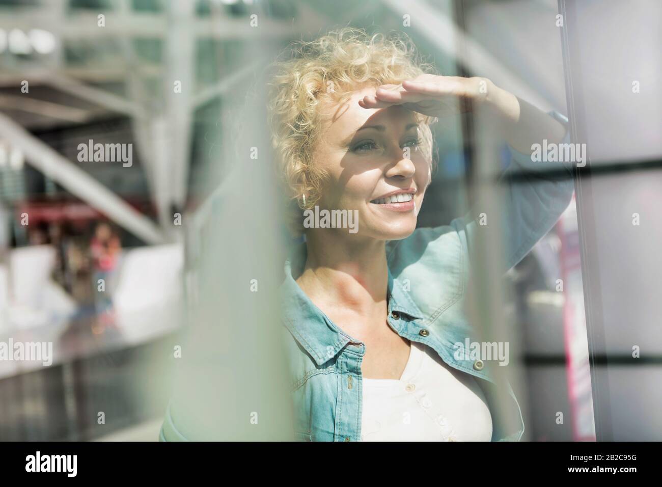 Porträt einer reifen Frau, die ihre Augen vor Sonnenlicht bedeckt, während sie durch das Fenster auf dem Flughafen blickt Stockfoto