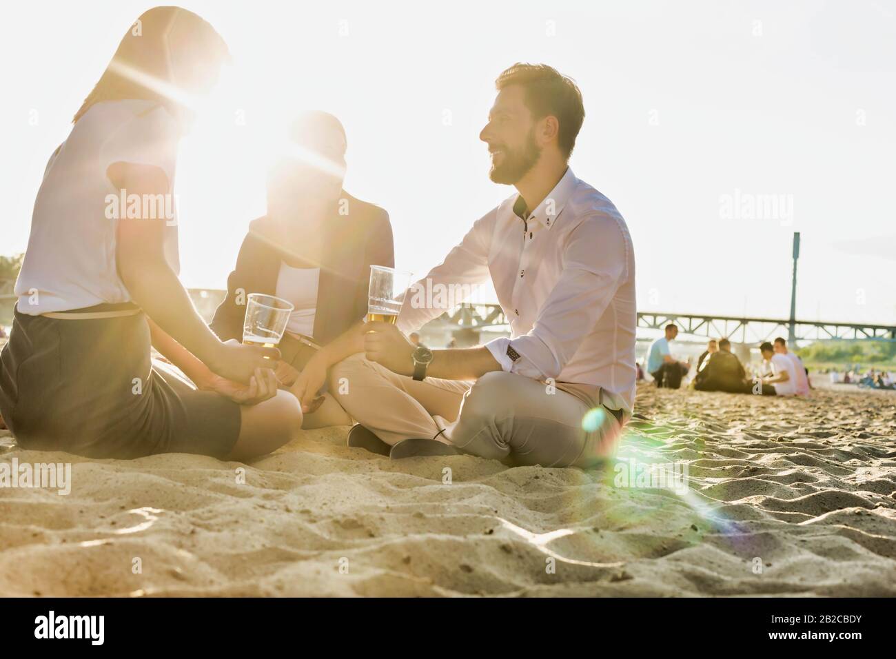 Geschäftsleute feiern Erfolg, während sie Bier am Strand mit Linsenflauge im Hintergrund trinken Stockfoto