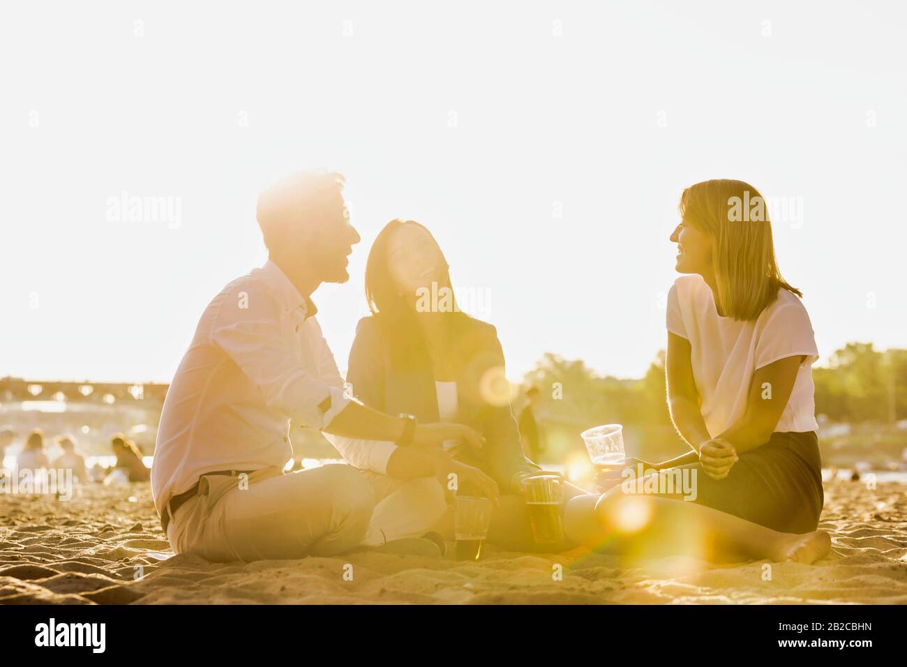 Geschäftsleute feiern Erfolg, während sie Bier am Strand mit Linsenflauge im Hintergrund trinken Stockfoto