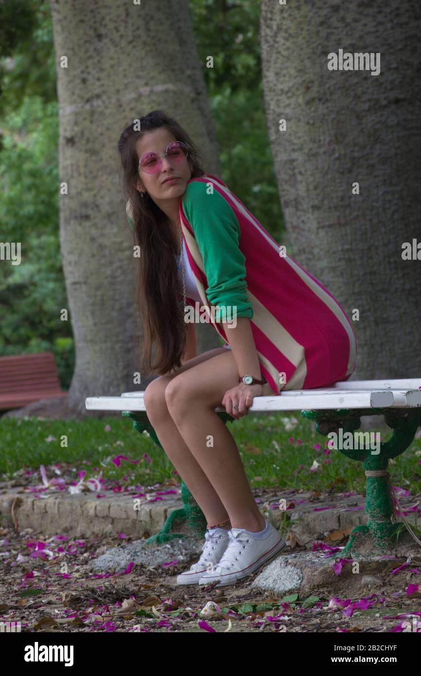 Nachdenkliche Frau, die auf einer Parkbank mit Kronblättern auf dem Boden sitzt Stockfoto