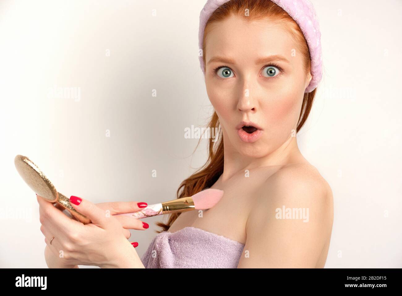 Ein rothaariges Mädchen in einem lilafarbenen Handtuch steht auf einem weißen Hintergrund, wobei ihre Augen überraschend offen sind und die Kamera betrachten. Stockfoto
