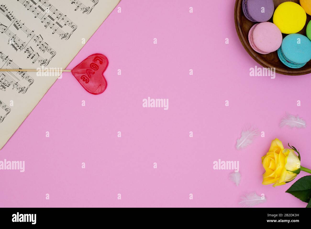 Rosa Hintergrund mit antiken Notenblättern, französisch Macarons Süßigkeiten, gelbe Rose, Herz geformt Lollipop Liebe und kleine weiße weiche Federn.Draufsicht. Für Stockfoto