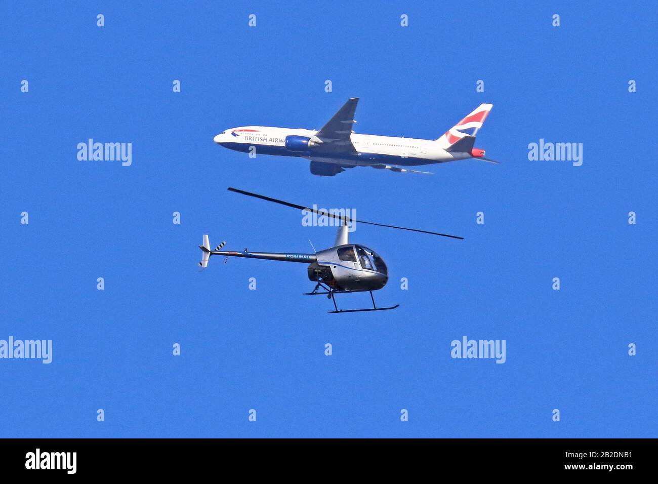 Perspektivische Verzerrung eines Robinson R22 Hubschraubers (G-DOGI) mit einer Boeing 777 der British Airways, die in entgegengesetzte Richtungen fliegt, gegen einen blauen Himmel gerichtet. Stockfoto