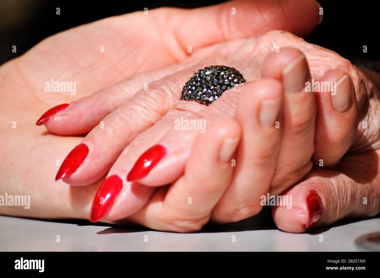 Erwachsene Tochter hält Mutter zerknitterte alte Hand, die einen Ring und einen roten nagellack trägt. Szene wurde teilweise mit einem dramatischen Softseffekt beleuchtet. Hände auf dem Tabl Stockfoto