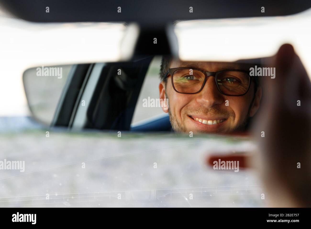 Fröhlicher fröhlicher Mann, der eine Brille trägt und Spiegel beim Sitzen in seinem Auto anpasst, sieht in Reflexion aus. Weicher Fokus. Emotionen beim Fahren eines neuen Autos Stockfoto