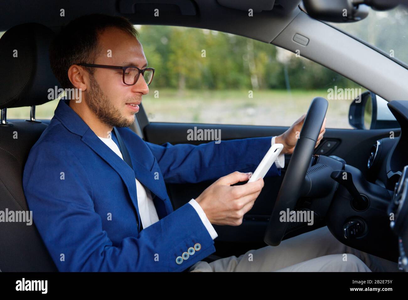 Seitenansicht eines lächelnden jungen Geschäftsmannes in einer Brille, die im Auto sitzt und auf dem Smartphone tippt/nachgibt, angehalten, um nicht abgelenkt zu werden. Stockfoto