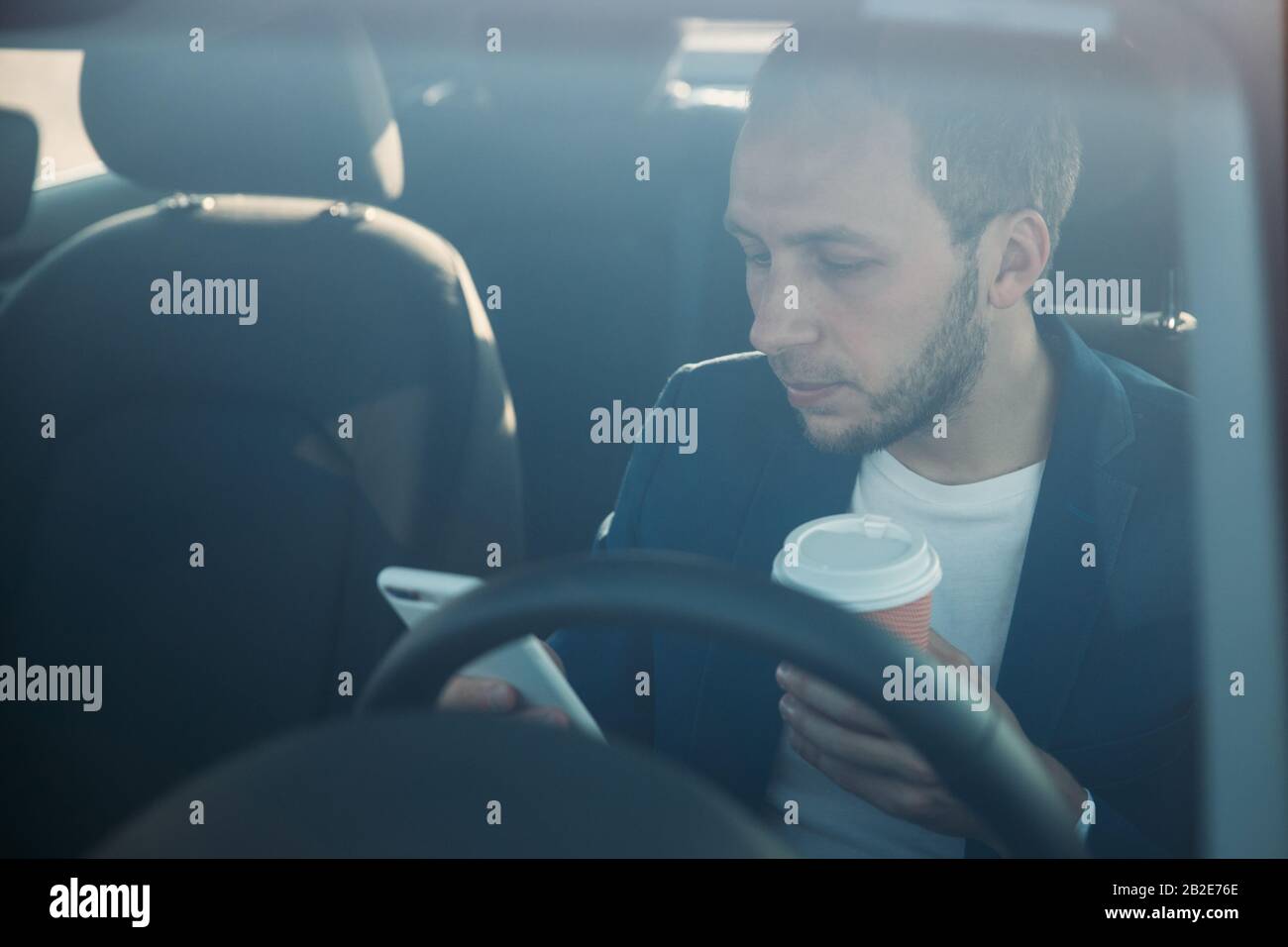 Vorderansicht des jungen Geschäftsmannes, der im Auto sitzt und auf dem Smartphone tippt/nachgibt, Kaffee in einer Papierbecher trinkt, durch die Windschutzscheibe gesehen. Kaffee B. Stockfoto