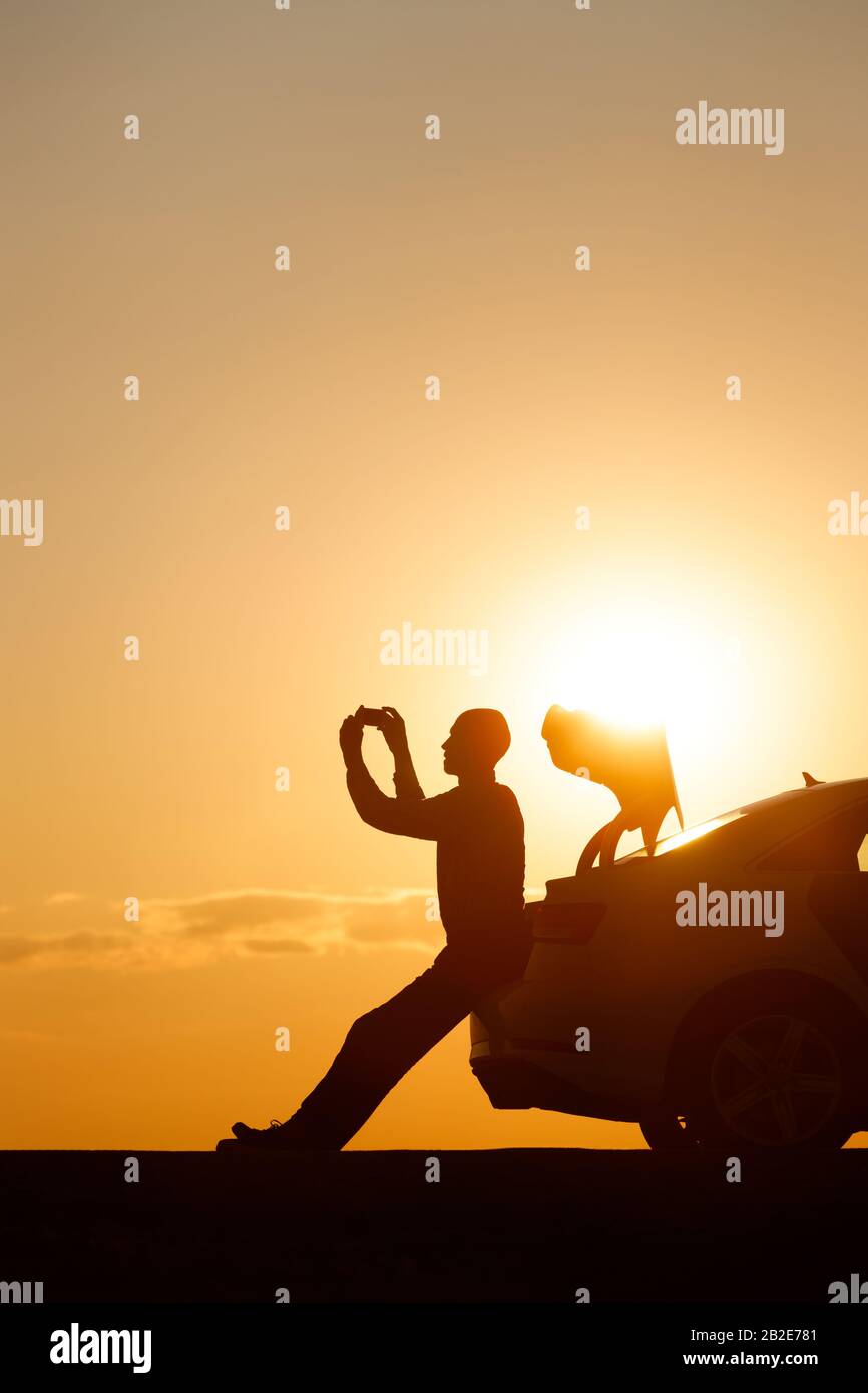 Silhouette eines Mannfahrers, der sich nach einer Fahrt entspannt, auf dem Kofferraum seines Autos sitzt und ein Foto zum Sonnenuntergang auf dem Smartphone, Seitenansicht, macht. Sommerberufung. Co Stockfoto