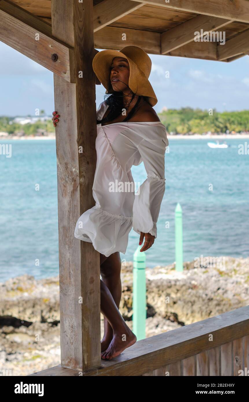 Schwarze Frau im Alter von 25 bis 30 Jahren, im Hafen sitzend, mit weißer Modelierung, frischer Sommer- und Tropenkleidung, mit Strandlandschaft und sonnigem Himmel Stockfoto