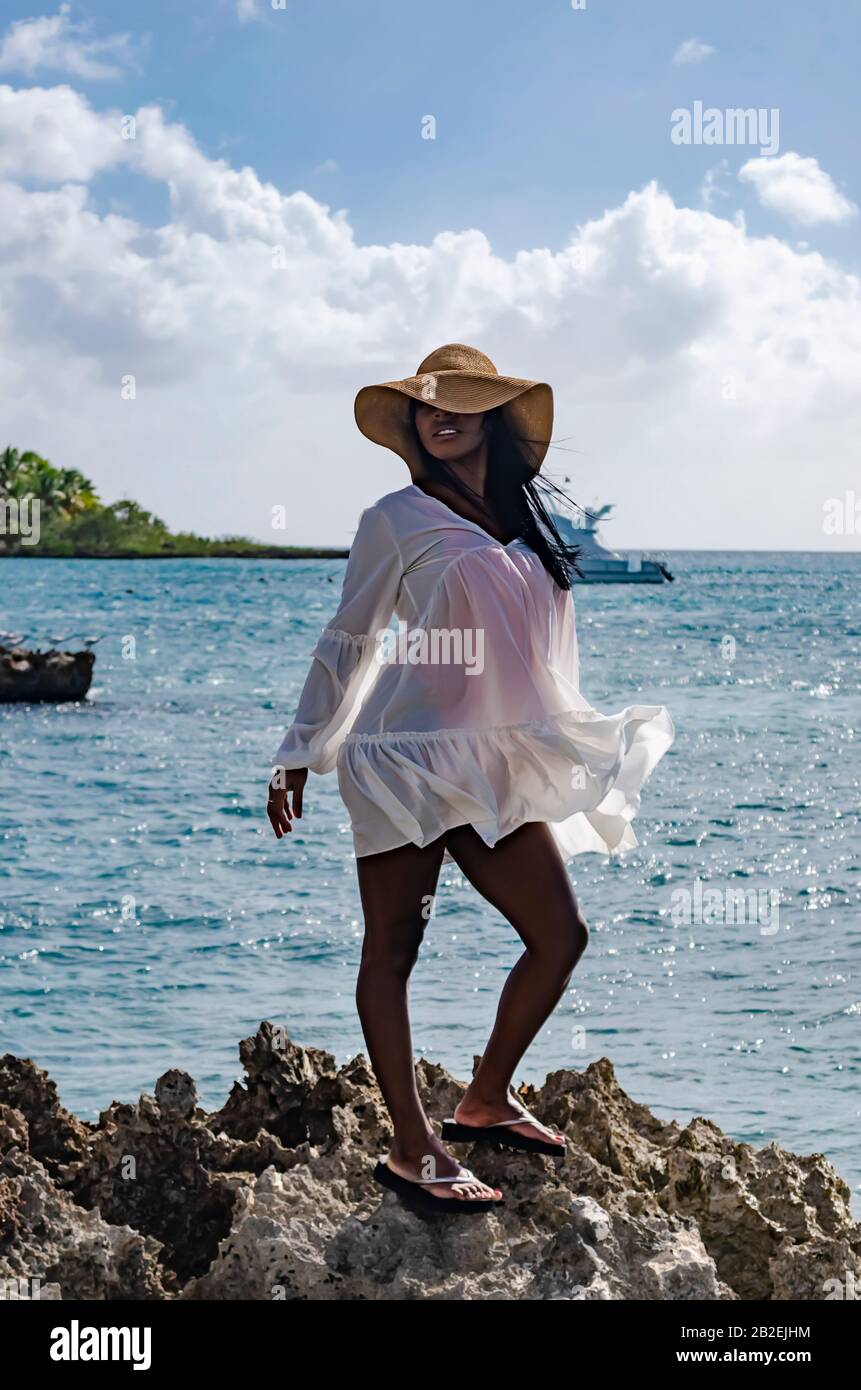 Schwarze Frau im Alter von 25 bis 30 Jahren, auf Felsen stehend, mit weißer Modelierung, frischer Sommer- und Tropenkleidung, mit Strandlandschaft und sonnigem s bekleidet Stockfoto