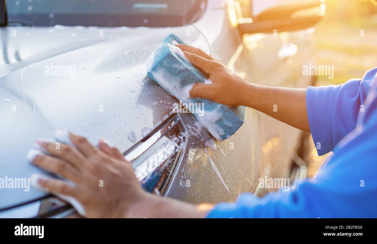 Asiatischer Mann mit blauem Schwamm und Seife zum Waschen des Autos im Freien bei Sonnenuntergang. Konzept für die Reinigung und Wartung von Autos Stockfoto