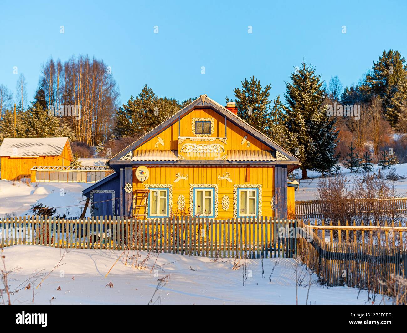 Winterblick auf Holzhaus im Dorf im russischen Norden. Altes Haus auf dem Land in wunderschöner Sonneneinstrahlung gegen einen blauen Himmel. Kaltes Wetter Stockfoto