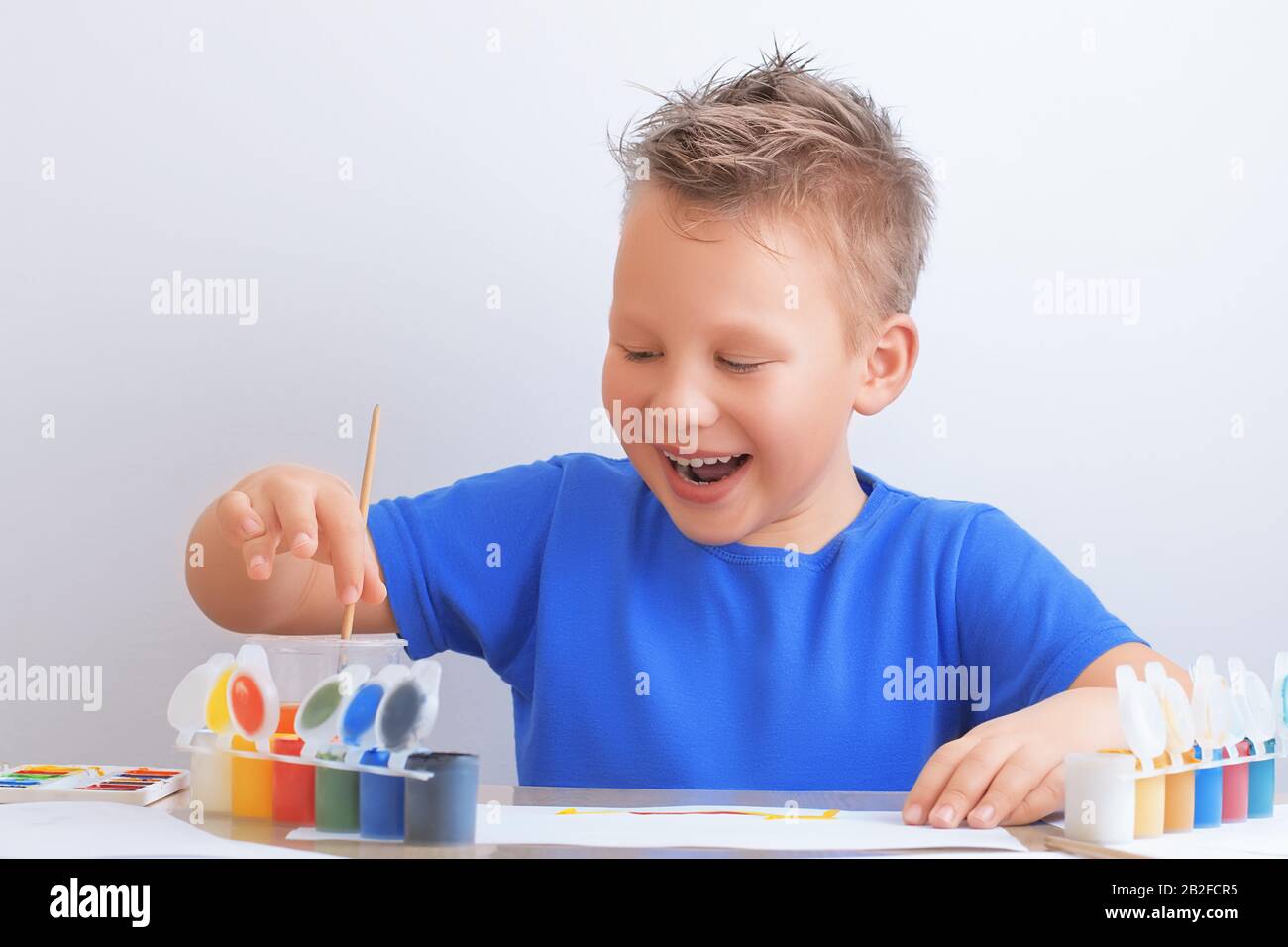 Lachender fröhlicher siebenjähriger Junge mit unordentlichen Haaren hält einen Pinsel in den Händen und zieht mit bunten Farben Stockfoto