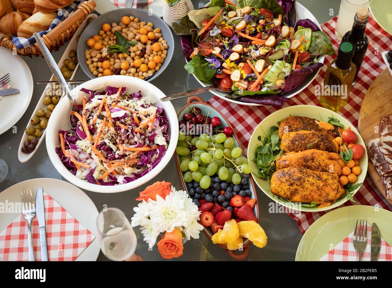 Blick über die Schüsseln mit Sommersalaten, gekochtem Fleisch und Obst, bereit zum Essen auf einem Tisch im Freien mit einem roten Gigham-Tischtuch, dekoriert mit Blumen. Stockfoto