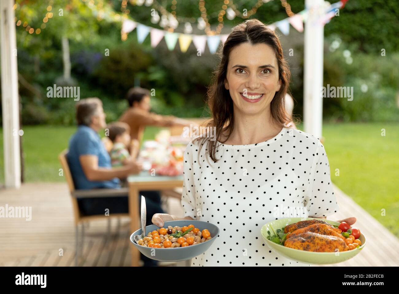Porträt einer lächelnden kaukasischen Frau, die Teller mit Essen hält, mit ihrer mehrköpfigen kaukasischen Familie, die draußen an einem Esstisch sitzt Stockfoto