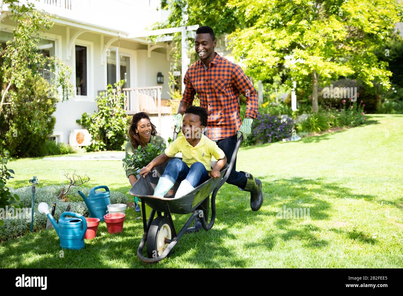 Vorderansicht eines glücklichen afroamerikanischen Paares und ihres Sohnes im Garten, der Mann schiebt seinen Sohn in einen Schubkarren, während die Mutter zuschaut. Familie e Stockfoto