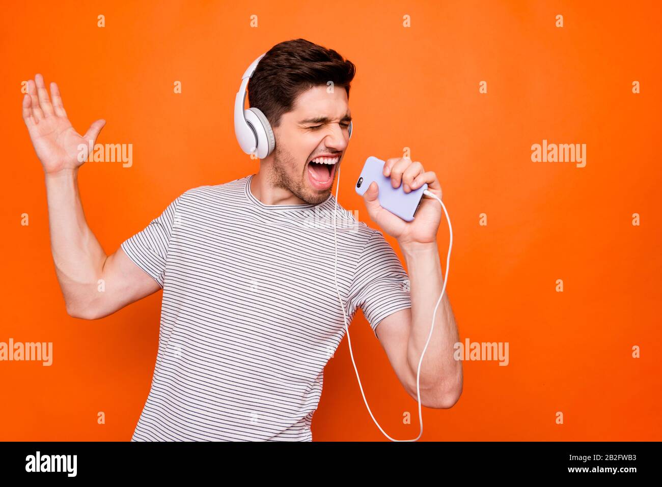 Foto von coolem Kerl fröhliche Party-Stimmung kühlende Hör-Ohrhörer halten Telefon wie Mikrofonaufnahme Single Wear gestreiftes T-Shirt isoliert Stockfoto