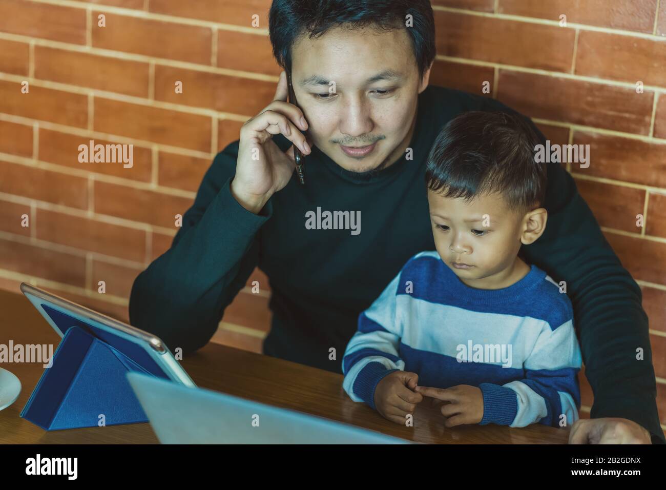 Asiatische alleinerziehende Väter mit Sohn sehen den Cartoon über einen Technologie-Laptop zusammen mit einem Telefon, das nach Arbeit ruft, wenn sie im Loft-Haus für Selbstlernende leben Stockfoto