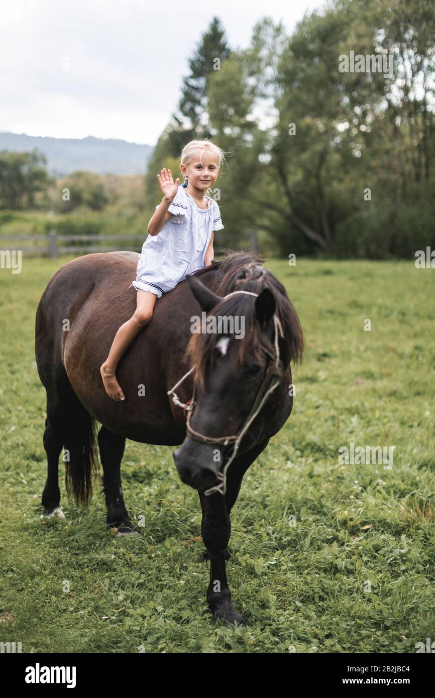Bild eines hübschen, blonden, haarigen Mädchens, das im Sommerfeld auf wunderschönem Pferd reitet, winkt und lächelt an die Kamera. Naturspaziergang, Reiten, Kinder und Stockfoto