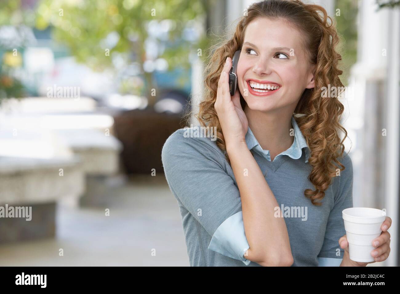 Junge Frau mit Getränk über Handy im Freien Stockfoto