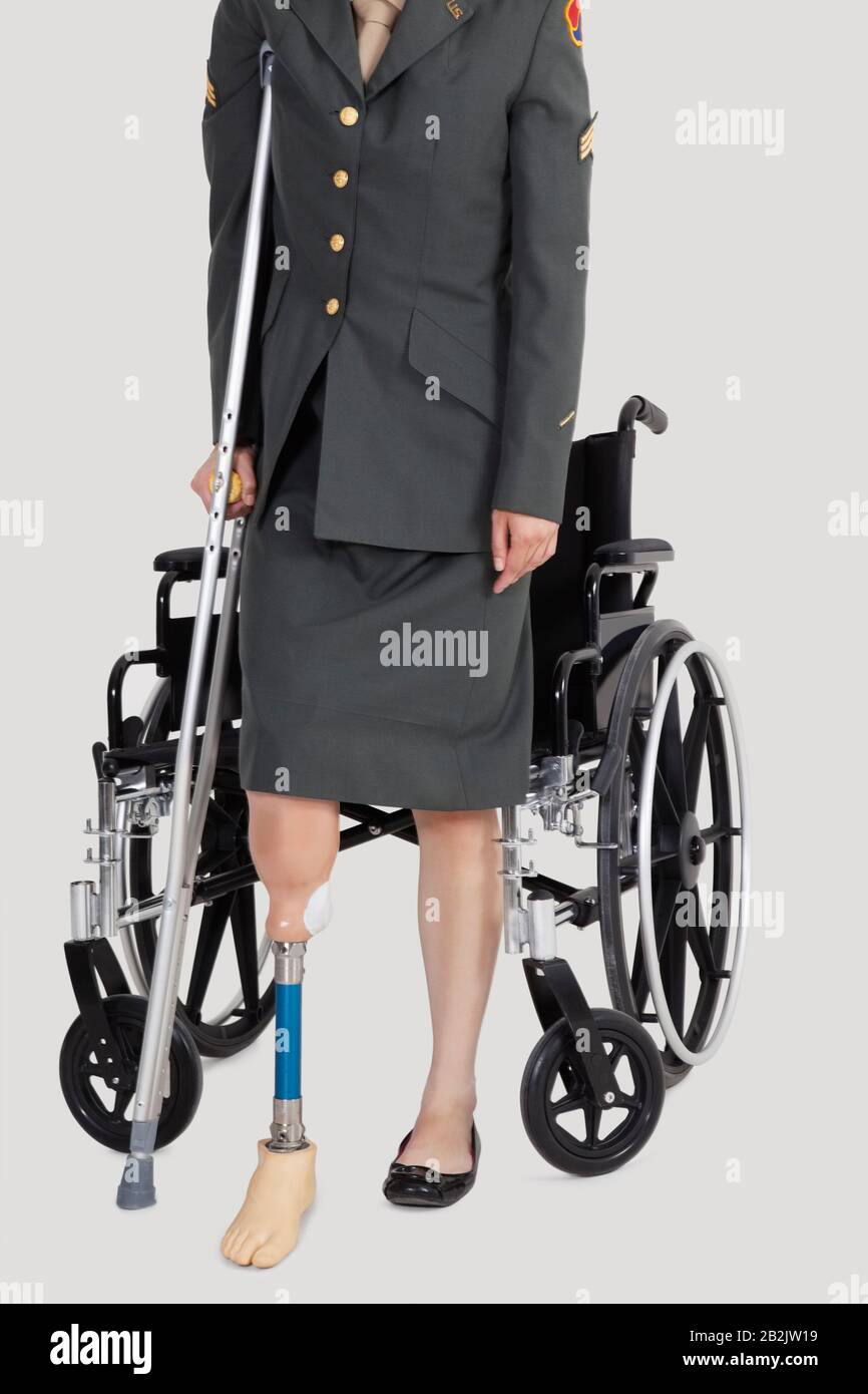 Niedriger Abschnitt eines weiblichen US-Militärbeamten mit Krücke, die vor dem Rollstuhl über grauem Hintergrund steht Stockfoto