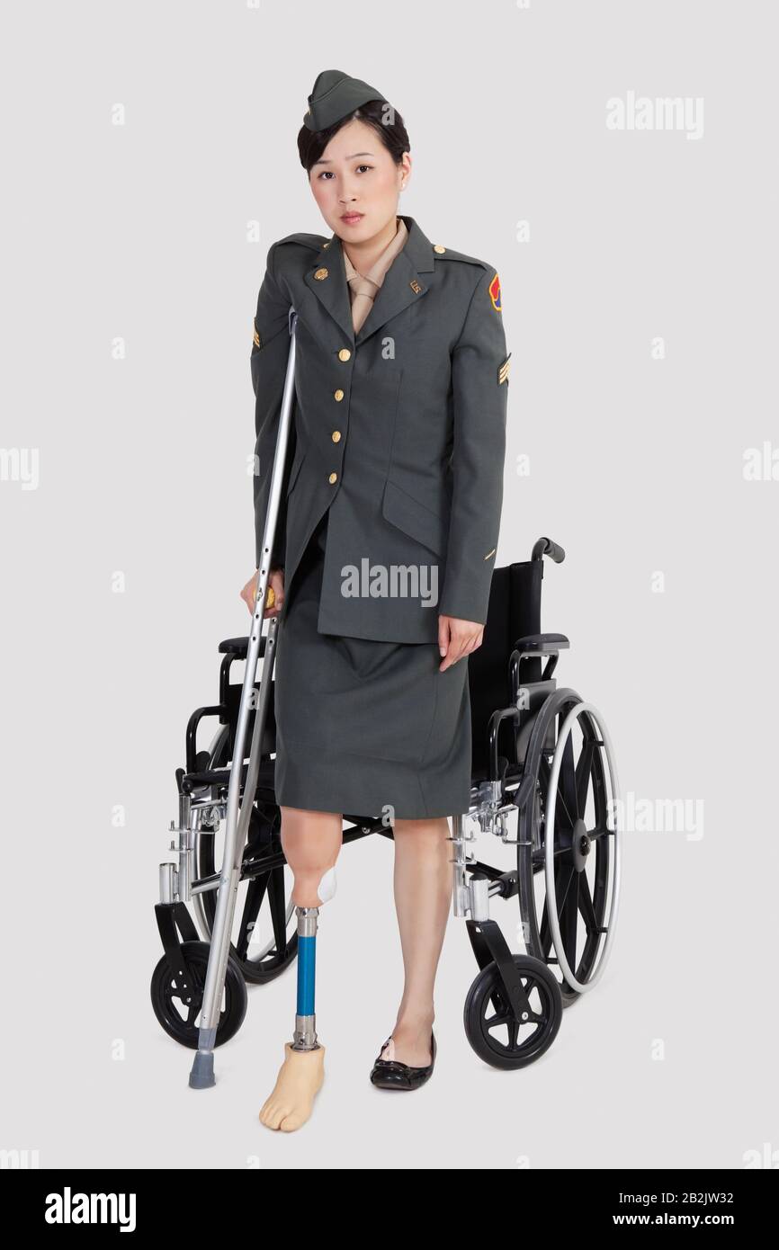 Behinderte US-Militäroffizierin mit Krücke, die vor dem Rollstuhl über grauem Hintergrund steht Stockfoto
