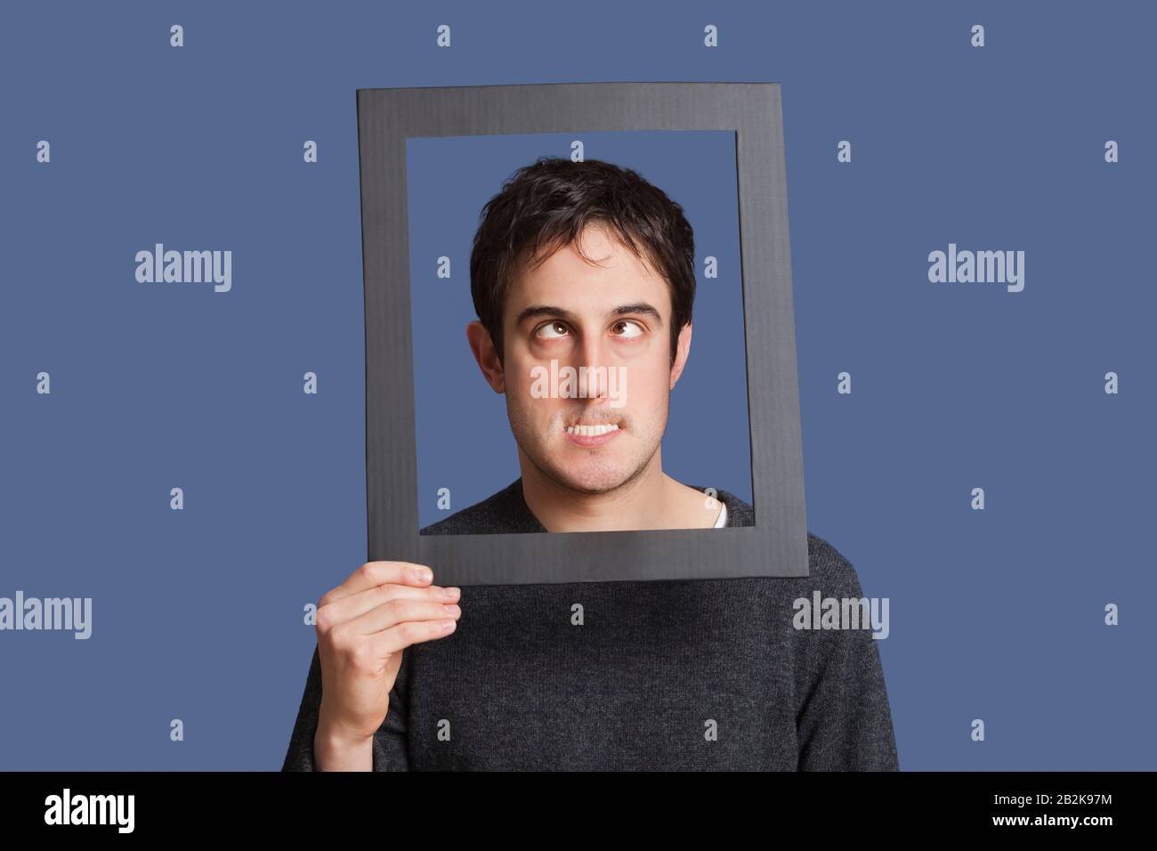 Junger Mann, der lustige Gesichter durch den Rahmen über blauem Hintergrund macht Stockfoto