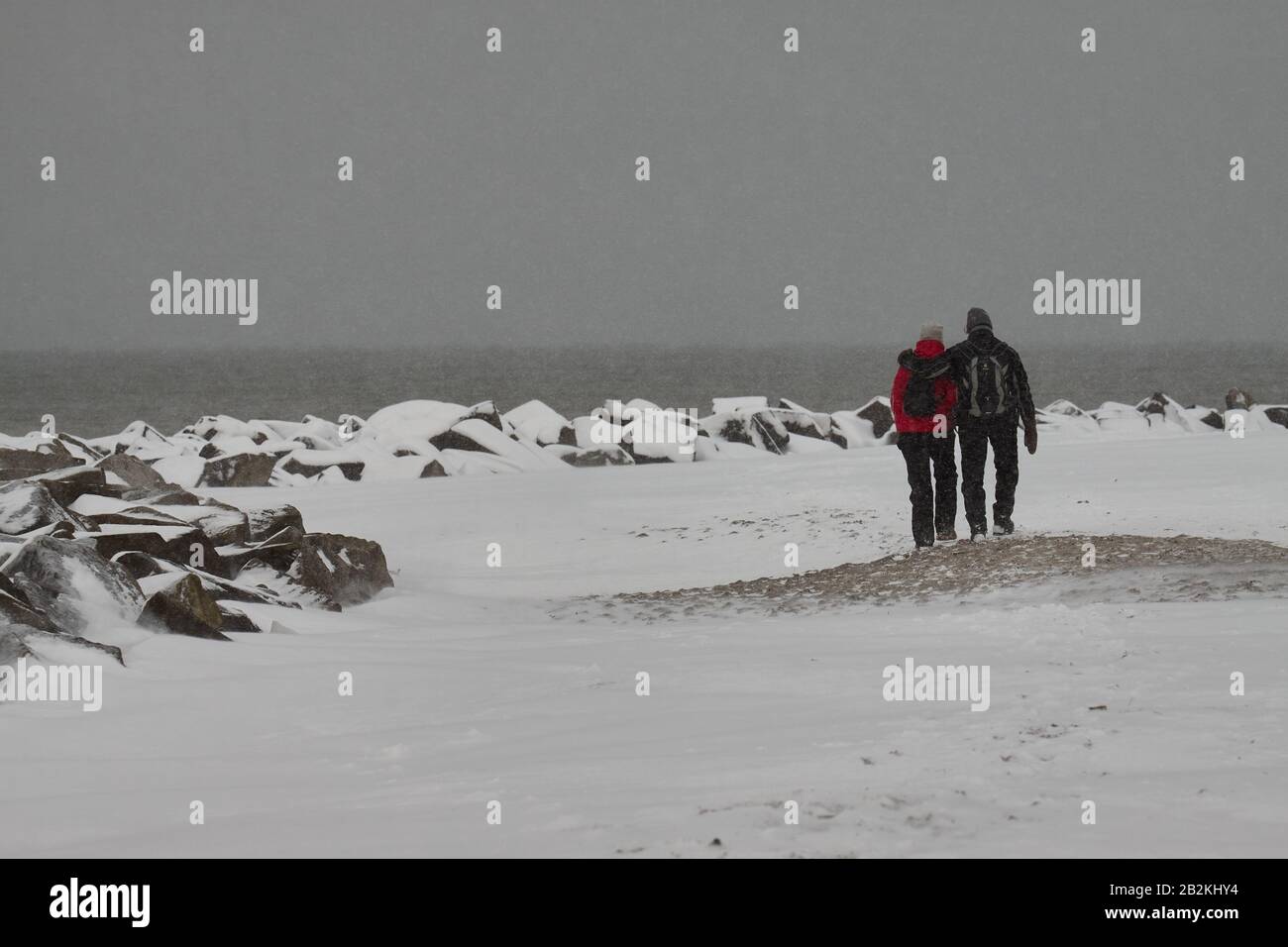 Unidentifizierbares Paar mit dem Rücken zum Kamer, der an einem verschneiten Strand mit schwarzen Felsen und schiefergrauem Himmel und Meer spaziert Stockfoto