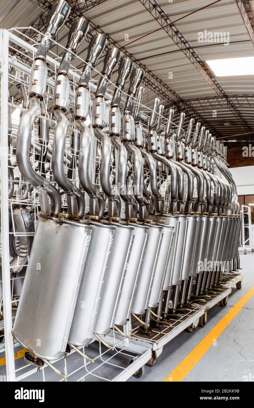 Mehrere Edelstahl-Auspuffanlagen, die jeweils aus einem gebogenen Rohr und einem zylindrischen Schalldämpfer bestehen, sind in ordentlichen Reihen auf weißen Metallregalen in einer Fabrik oder einem Lager angeordnet. Die Racks sind Stockfoto