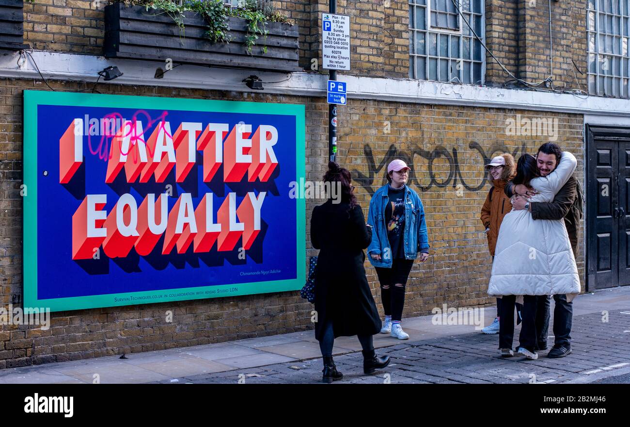 Eine Gruppe junger Leute trifft sich und umarmt und lächelt vor einer großen Graffiti-Art-Plakatwand, die besagt, dass ich In der Brick Lane in East London Gleich Wichtig bin. Stockfoto