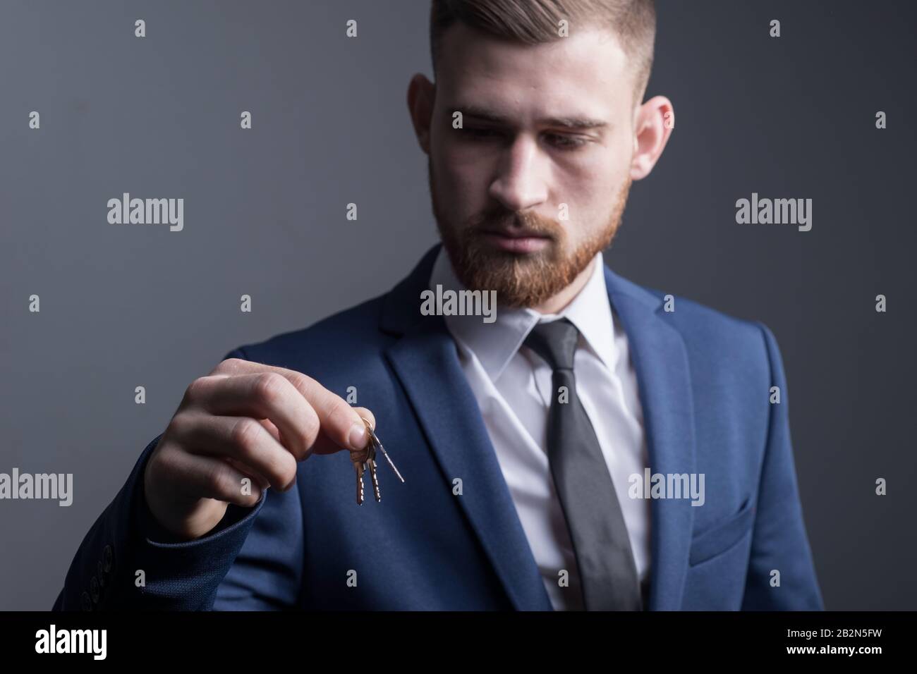 Nahaufnahme eines jungen bärtigen, fünfundzwanzigjährigen Mannes in einem Business-Anzug, der Schlüssel in der Hand hält. Auf grauem Hintergrund. Konzept. Käuflich Stockfoto