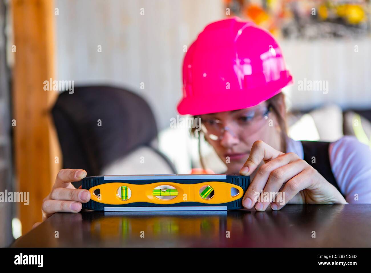 Nahaufnahme und selektive Fokusaufnahme des von einer Architektin verwendeten Spirit Level Werkzeugs, sie trägt im Hintergrund eine rosafarbene Hardhut und eine Schutzbrille Stockfoto