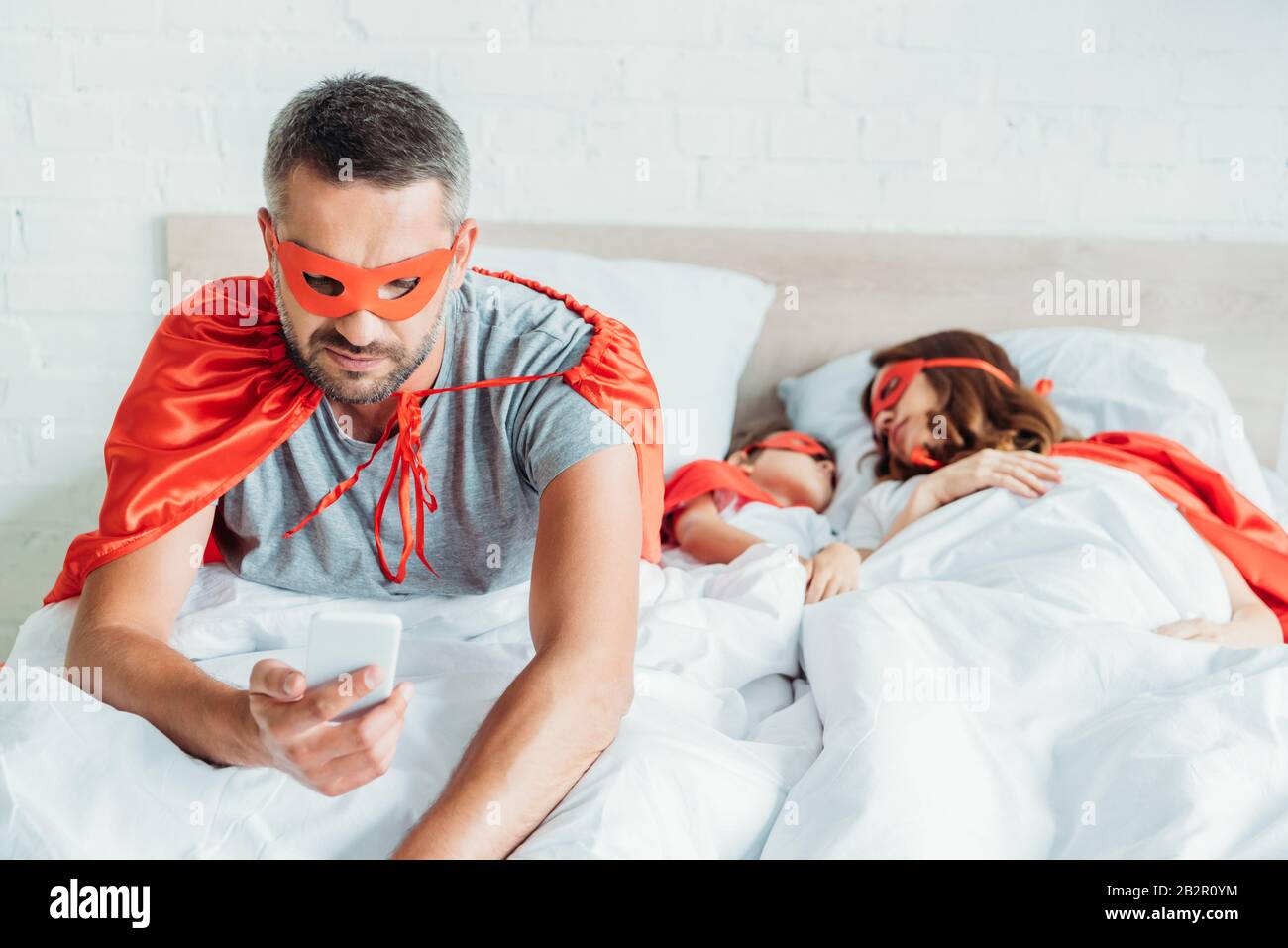 Mann im Kostüm des Superhelden, der Smartphone benutzt, während er im Bett  in der Nähe einer schlafenden Familie sitzt Stockfotografie - Alamy