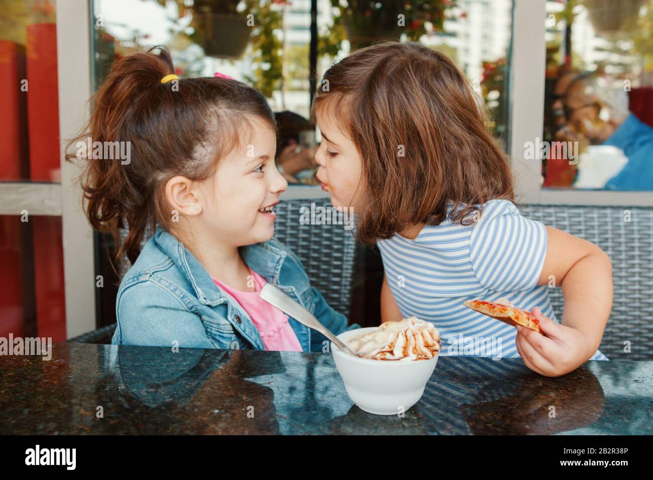 Zwei lustige kaukasische kleine Vorschulschwestern küssen sich im Café. Freunde Mädchen haben Spaß zusammen. Kinder, die essen, frühstücken auf der Terrasse des Restaurants. Stockfoto