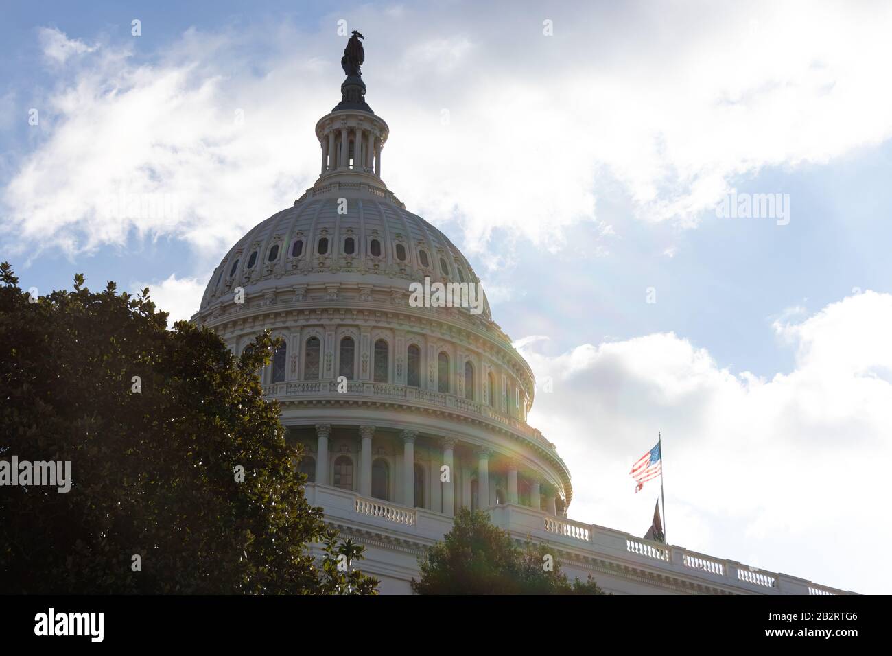 United States Capital Dome hinter Bäumen an einem sonnigen Nachmittag, schwenkt die amerikanische Flagge in den Wind. Stockfoto