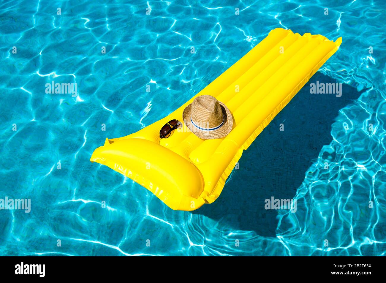Seine und seine Sonnenbrille ruhen auf leerem, gelb aufblasbarem Floß, das im hellblauen Schwimmbad schwimmt Stockfoto