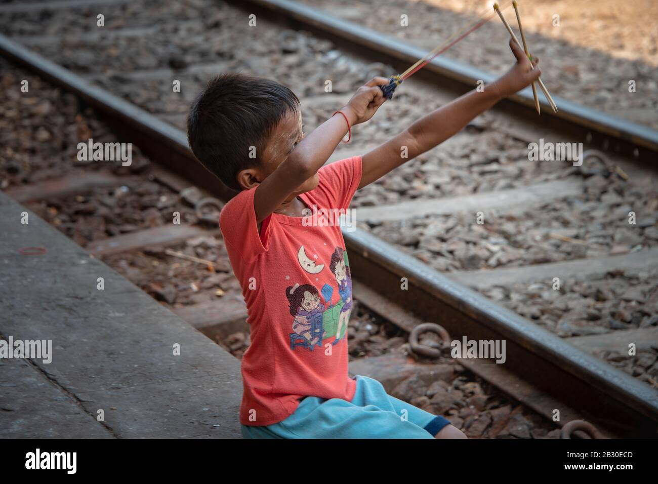 Yangon, Myanmar - Januar 2020: Junger asiatischer Junge, der mit einem einfachen hausgemachten Katapult neben Bahngleisen spielt, Stockfoto