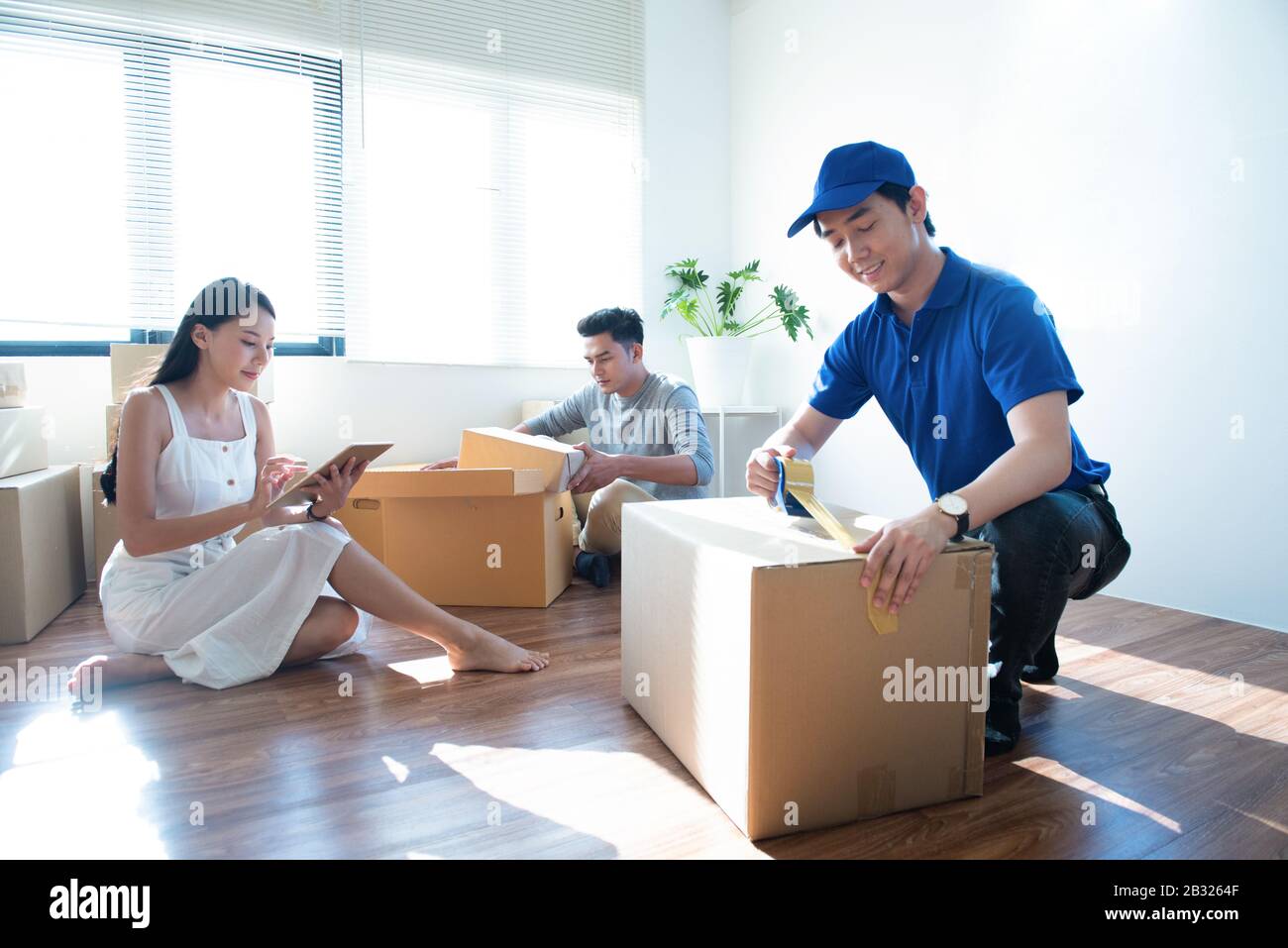 Der Mann des Lieferdienstes in blauer Uniform hilft dem glücklichen Paar Kunden, das Paket mit Klebeband im Haus des Kunden verpackt.Frau mit Tablettenunterschrift Stockfoto