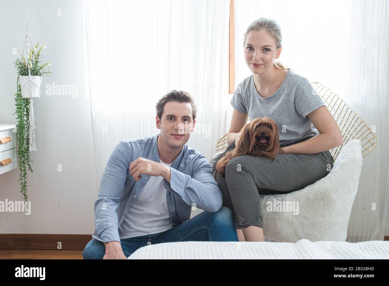 Junges glückliches kaukasisches Paar mit Lächeln sitzt auf dem Sofa und umarmt den Hund am Knie im Schlafzimmer zu Hause, im Innenbereich Lifestyle-Konzept. Stockfoto