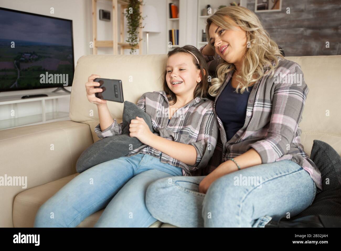 Mutter und Tochter mit passender Kleidung, die auf der Couch sitzt und ein lustiges Video auf dem Smartphone sieht. Stockfoto