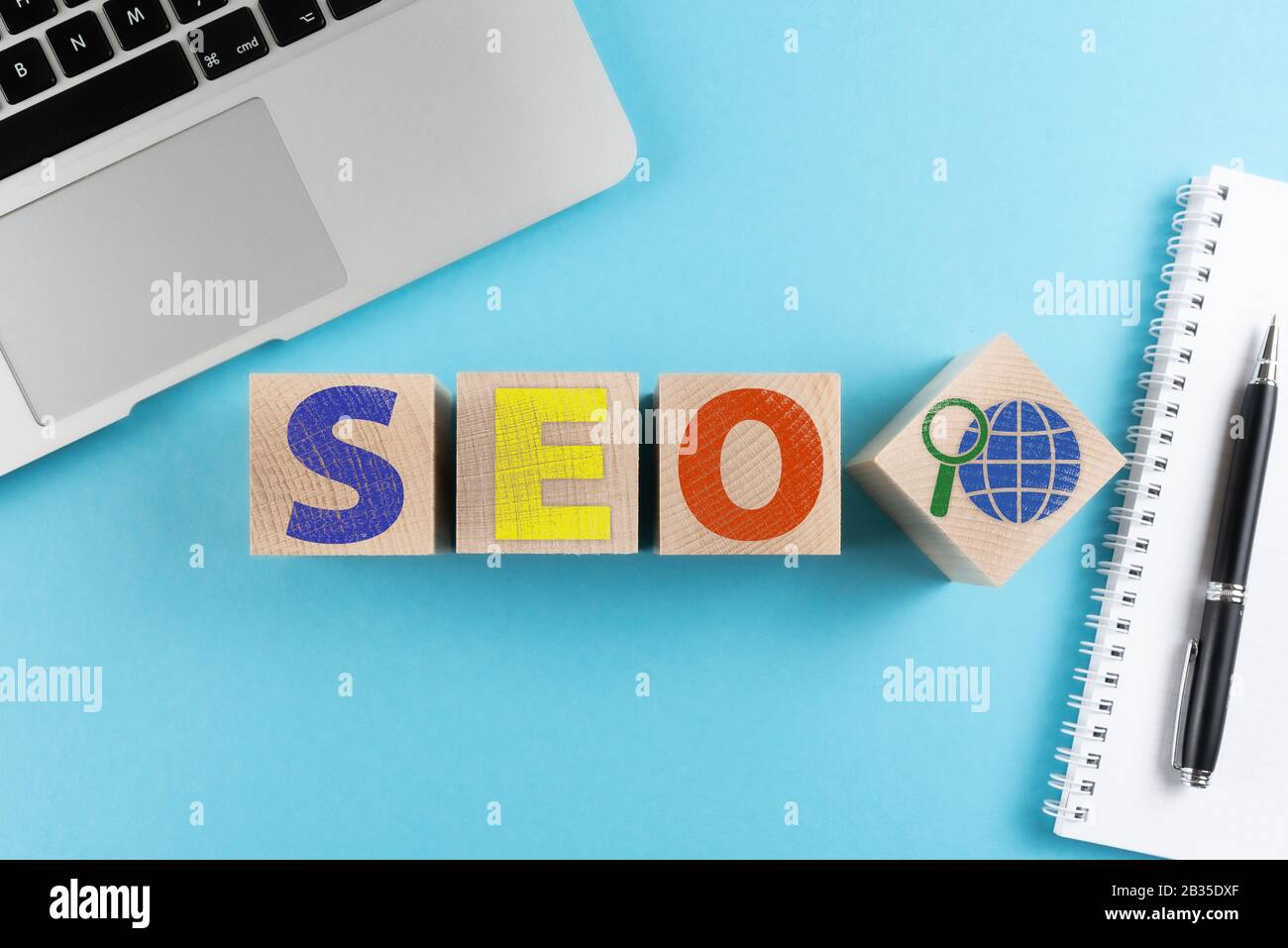 Draufsicht über die Buchstaben SEO und Internetsuchsymbol auf Holzwürfeln auf blauem Hintergrund mit Laptop und Spiralnotizbuch Stockfoto