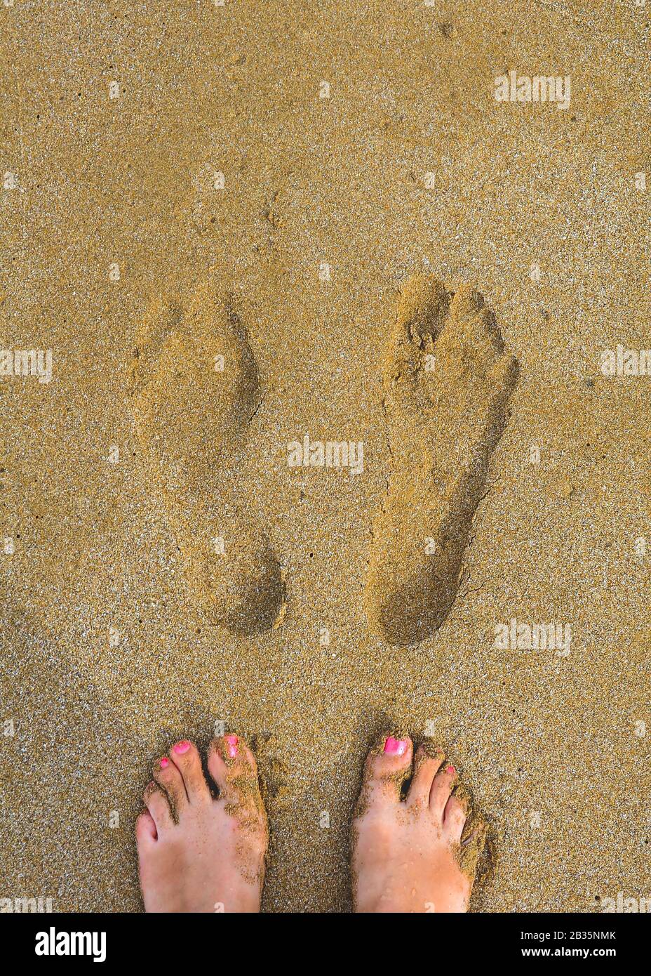 Barfüßige weibliche Füße mit rotem Pediküre neben ihren eigenen Drucken auf dem nassen gelben Sand. Draufsicht. Stockfoto