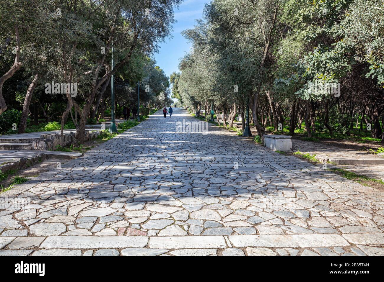 Gepflasterter Fußweg aus altem Marmorstein am Phiilopappou-Hügel in Athen, Griechenland. Die Menschen gehen an einem sonnigen Tag einen Kopfsteinpflaster unter der alten Akropolis. Bäume a Stockfoto