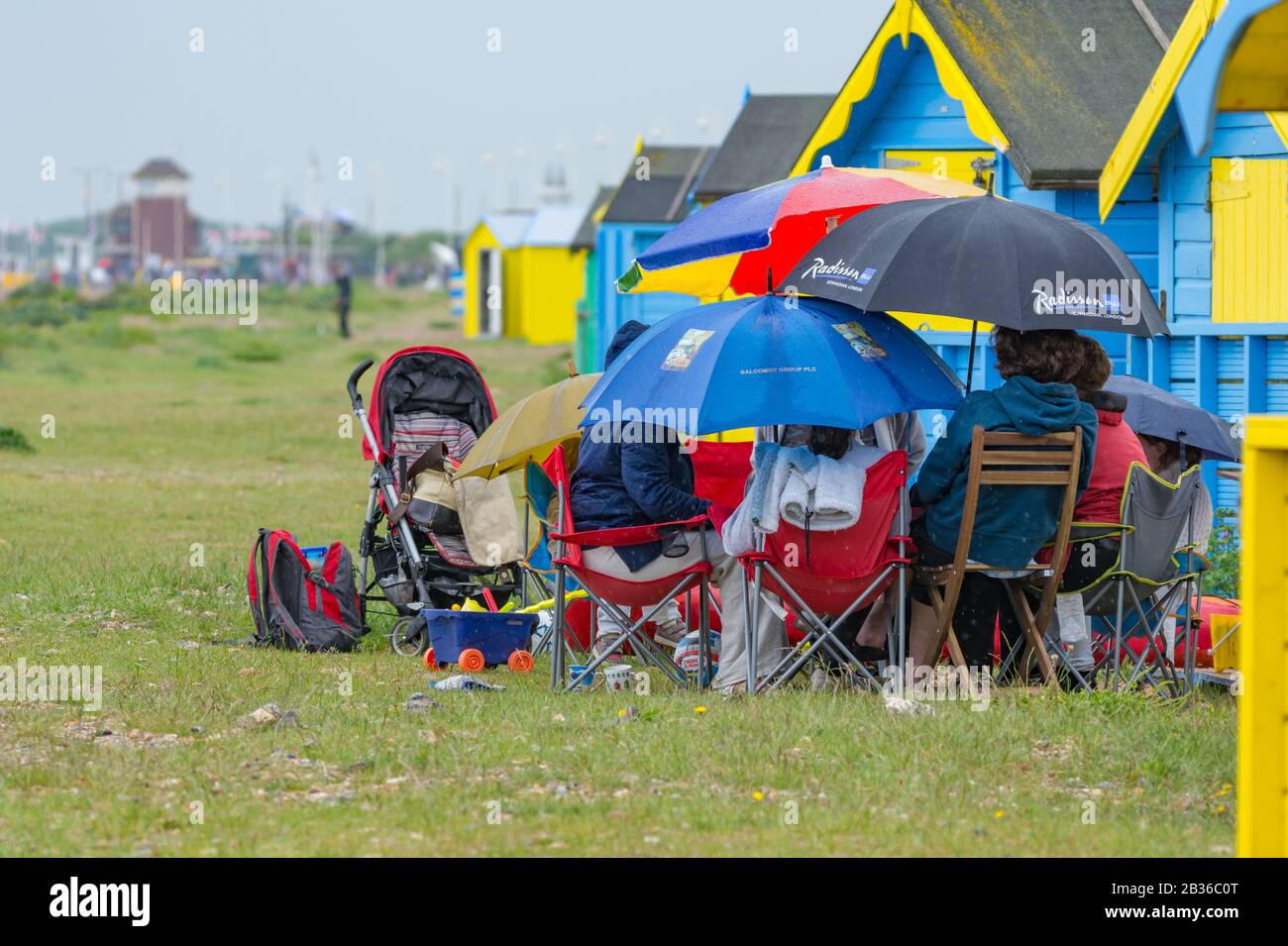 Leute, die draußen im Regen auf einem Strand sitzen, der Regenschirme hält, an einem nassen elenden regenden Bankfeiertag am Montag an einem typischen britischen Wettertag, Großbritannien. Stockfoto
