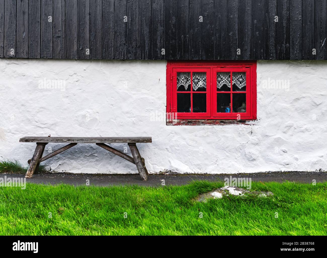 Weiße Hauswand mit rotem Fenster auf altem färöischem Haus. Holzbank auf grünem Gras. Faröer Inseln, Dänemark Stockfoto